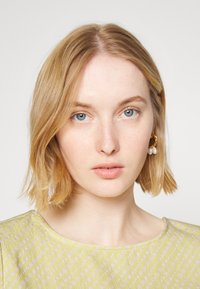 Cabelo louro e olhos azuis. Design de gola redonda em tecido verde claro com pequenos pontos rosa pálido. Brincos de argola dourados com detalhes em pérola.