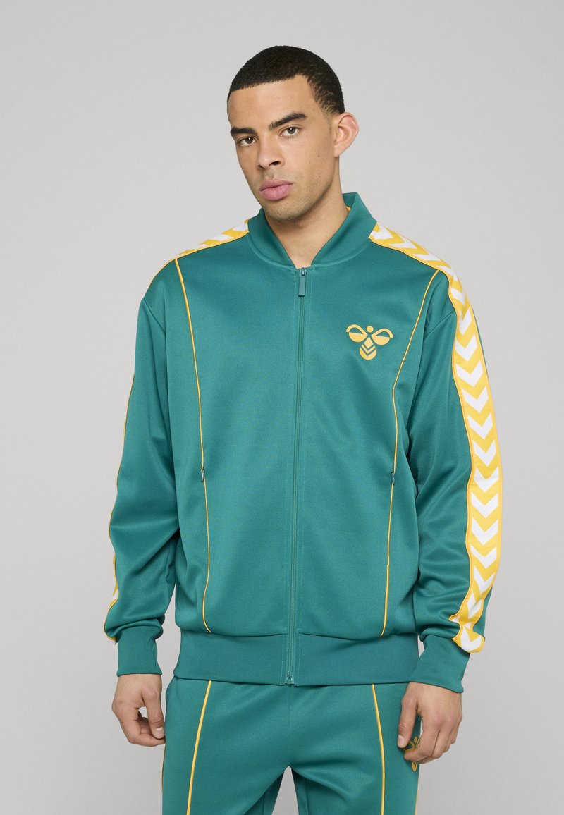 Veste de sport bleu sarcelle avec une fermeture éclair intégrale, des accents jaunes et un logo d'abeille. Comprend des bandes à motif chevron sur les manches et des poignets côtelés.