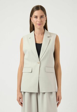 Frau mit langen braunen Haaren trägt einen ärmellosen beigen Blazer über einem schwarzen Top und passenden beigen, weit geschnittenen Hosen, schaut nach vorne vor weißem Hintergrund.