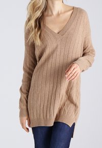 Femme portant un pull beige côtelé à col en V avec manches longues et fentes sur les côtés, associé à un jean bleu foncé.