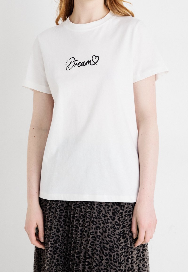 T-shirt en coton blanc avec du texte en cursive noir "Rêve" accompagné d'un cœur. Manches courtes et coupe décontractée. Porté par-dessus une jupe à imprimé léopard.