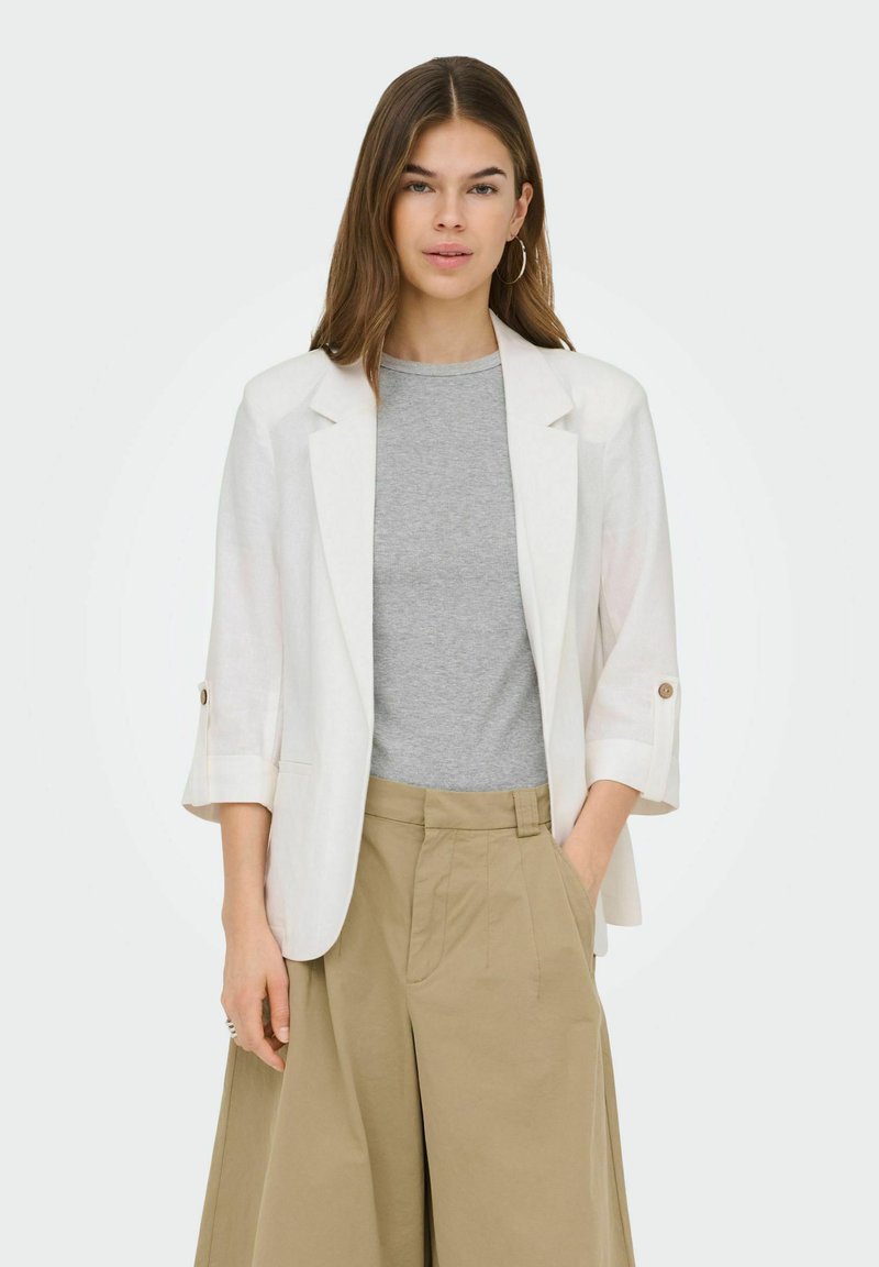 Femme portant un blazer blanc aux manches retroussées, un t-shirt gris col rond, un pantalon beige taille haute à jambes larges et des boucles d'oreilles créoles.