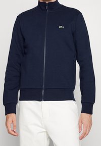Giacca con zip blu navy in tessuto morbido, caratterizzata da collo alto, polsini a coste e piccolo logo ricamato sul petto.