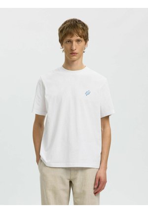 Młody mężczyzna w gładkiej białej koszulce z małym niebieskim abstrakcyjnym logo, beżowe spodnie, stojący na tle jednolitego jasnoszarego tła.