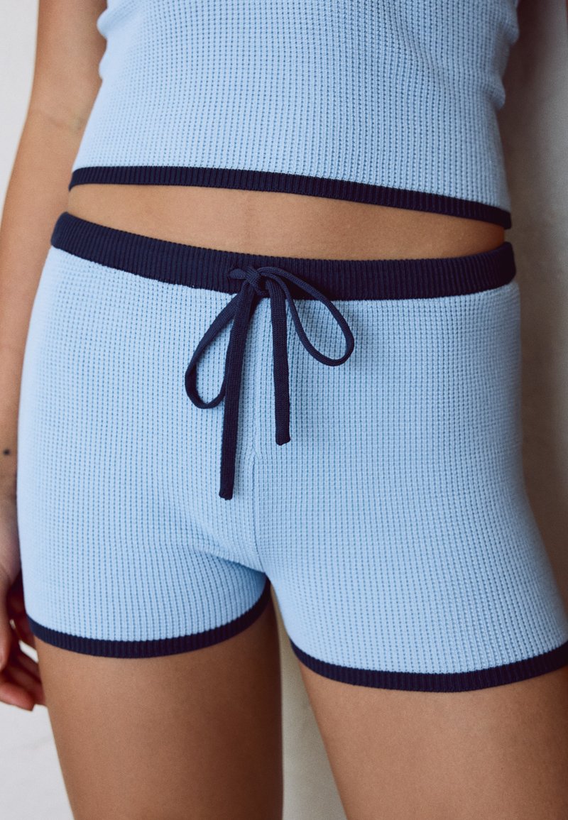Gros plan d'une personne portant un short en maille bleu clair avec une bordure bleu foncé et un cordon de serrage, associé à un haut sans manches en maille assorti.