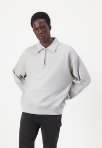 Sweatshirt cinza claro com gola com fecho, corte oversized e ombros caídos, combinado com calças pretas. Textura suave e costuras robustas.