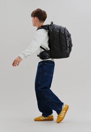 Junge, der in weißem Sweatshirt, blauer Jeans, gelben Turnschuhen und mit einem großen schwarzen Rucksack mit gemustertem Stoff geht.