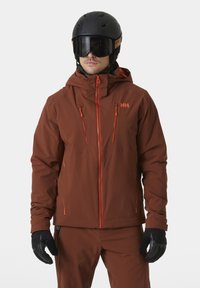 Hnědá zateplená lyžařská bunda s oranžovými zipy, kapucí a logem. Černá helma a rukavice doplňují design. Hladká textura po celém povrchu.