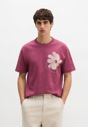 Junger Mann mit dunklem Haar, der ein mauvefarbenes T-Shirt mit weißem abstraktem Blumenmotiv und beige Hose trägt, steht mit den Händen in den Taschen.