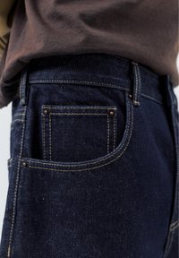 Zbliżenie na kieszeń ciemnoniebieskich dżinsów ze widocznym żółtym przeszyciem oraz częścią wsuniętej brązowej koszuli.