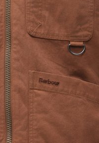 Prim-plan al unei jachete din țesătură maro, cu fermoar metalic, buzunare cusute, un inel metalic și logo-ul brodat "Barbour".