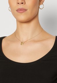 Collar de oro con un colgante rectangular texturizado, llevado en un cuello desnudo, combinado con una blusa negra de escote en barco. Diseño simple y elegante.
