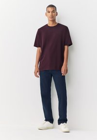 T-shirt di cotone color marrone con maniche corte e scollo rotondo, abbinata a pantaloni blu scuro e scarpe da ginnastica bianche, in piedi davanti a uno sfondo bianco.