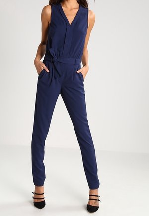 Granatowy kombinezon z dekoltem w serek, bez rękawów, z paskiem w talii i dwoma przednimi kieszeniami. Gładka tkanina z nogawkami zwężanymi ku dołowi.