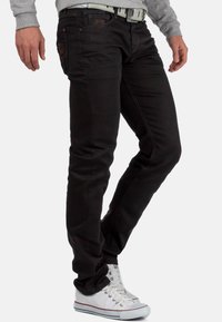Jean en denim noir avec une coupe slim, présentant des détails de couture subtils, cinq poches et une ceinture contrastante. Porté avec des baskets blanches.