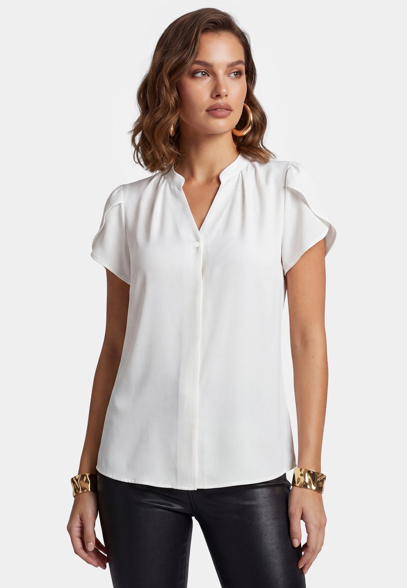 Femme aux cheveux bruns ondulés porte un chemisier blanc à manches courtes, un pantalon noir, de grandes boucles d’oreilles dorées en anneau et des bracelets manchettes en or, regardant vers la droite.