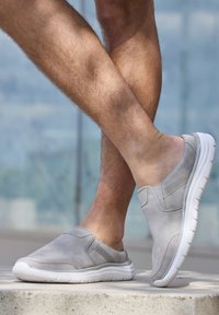 Gray slip-on shoes with white soles, featuring smooth suede material, stitching details, and elastic side panels. Displayed on crossed legs.