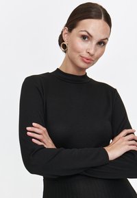 Czarny top z długim rękawem o stójce, wykonany z prążkowanego materiału, o dopasowanym kroju i gładkiej strukturze. Modelka pozuje z założonymi rękami.
