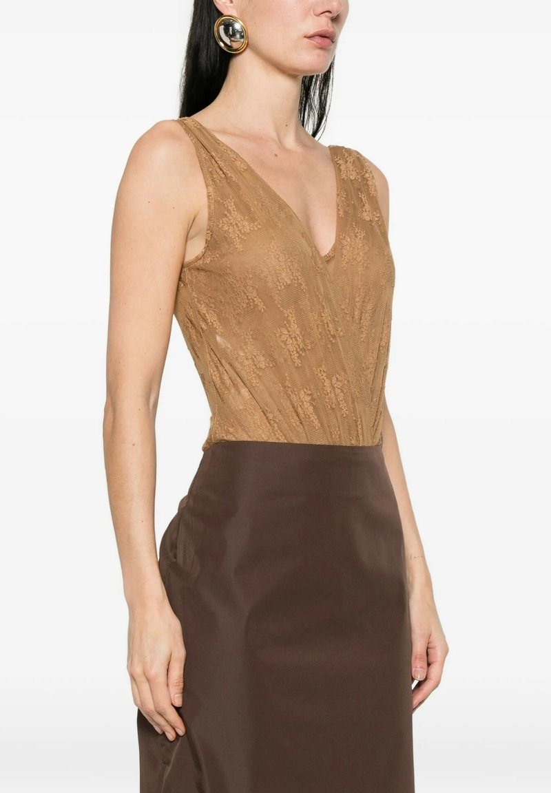 Donna che indossa un top smanicato color beige in pizzo con scollatura a V profonda e gonna a vita alta di colore marrone scuro, accessoriata con grandi orecchini rotondi dorati.