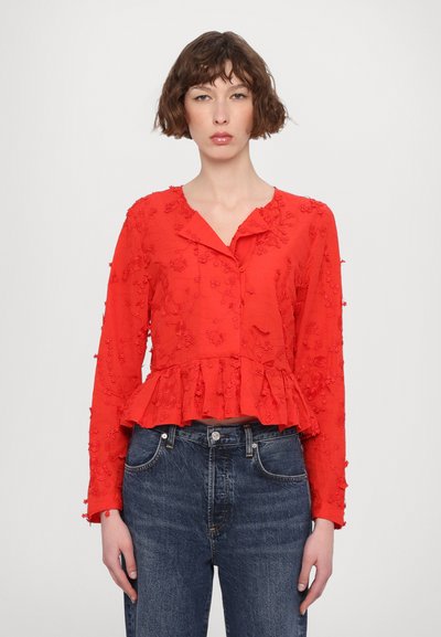 Rød bluse med blomsterbroderi, kravehals, lange ærmer og peplum-nederdel, parret med mørkeblå jeans.