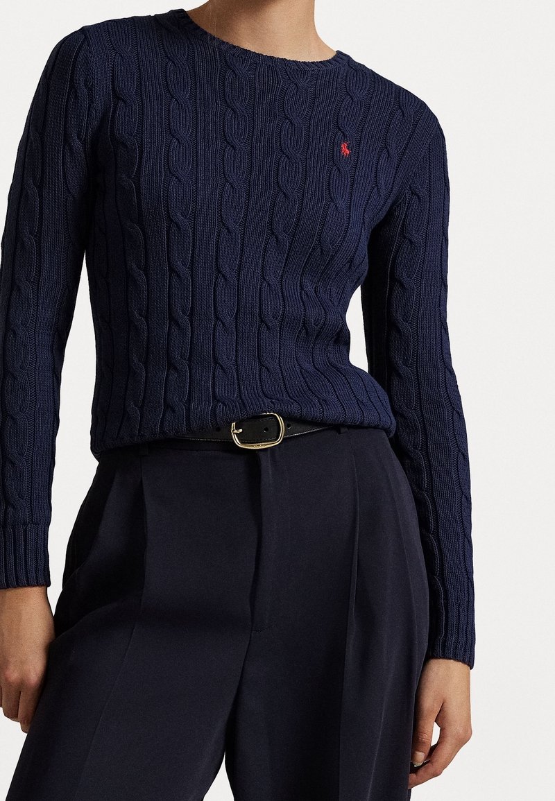 Personne portant un pull en maille torsadée bleu marine avec un petit logo rouge, associé à un pantalon noir taille haute et une ceinture noire avec une boucle dorée.