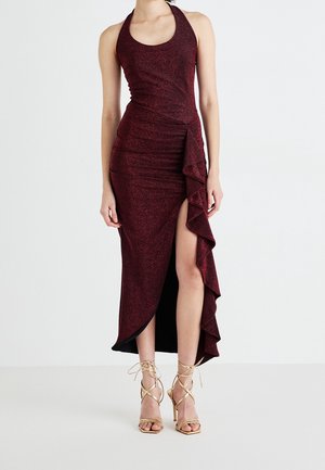 Kobieta ubrana w bordową sukienkę na halter bez rękawów z wysokim rozcięciem i falbaną, w połączeniu ze złotymi sandałami na obcasie z wiązaniem.