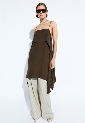 Donna che indossa un top drappeggiato senza maniche marrone scuro, pantaloni larghi chiari, scarpe bianche a punta e grandi occhiali da sole scuri, in piedi su sfondo bianco.