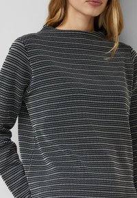 Czarnobiały wzorzysty top o wysokim dekolcie, z długimi rękawami i teksturowaną tkaniną z małymi wzorami przypominającymi siatkę.