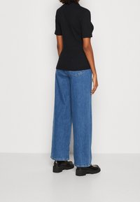 Kvinna som bär en svart kortärmad ribbad topp, lösa blå breda jeans och svarta skor med tjock sula, stående mot en ljus bakgrund.