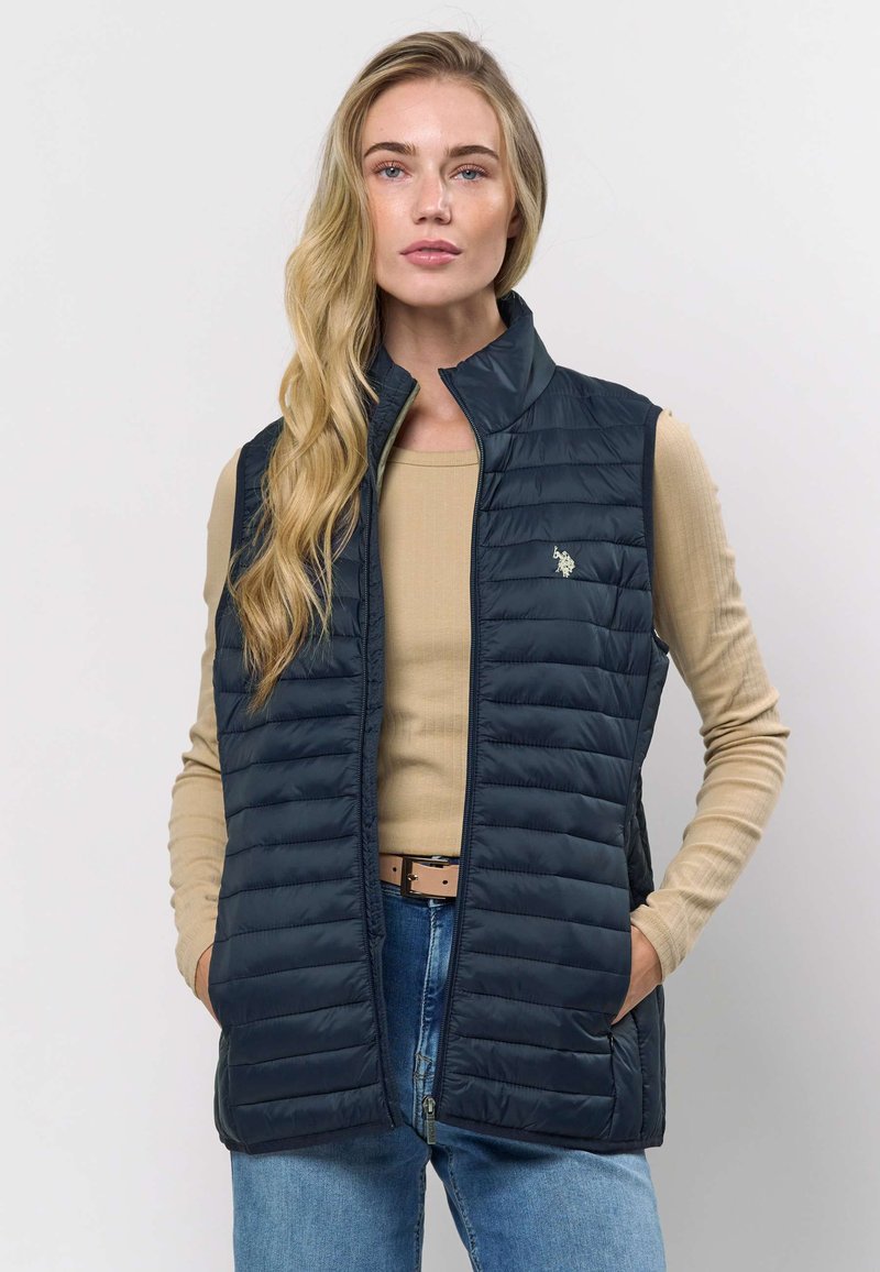 Mornarsko moder brezrokavnik z visokim ovratnikom, sprednjo zadrgo in bočnimi žepi. Nošen čez svetlo bež majico z majhnim logotipom na prsih.