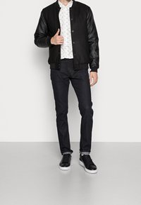 Giacca bomber nera con maniche in pelle, camicia bianca a fantasia, jeans scuri e sneakers nere. Design semplice e sportivo con texture a contrasto.