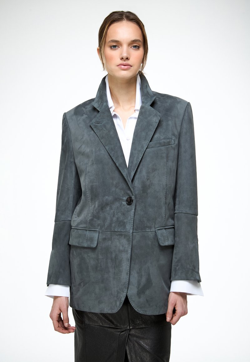 Joven mujer con un blazer de ante gris sobre una camisa blanca y una falda de cuero negra, posando delante de un fondo blanco liso.