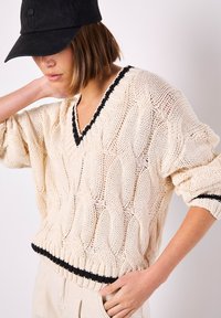 Beige Strickpullover mit V-Ausschnitt, schwarzem Saum am Kragen und an der Unterseite sowie lockeren Ärmeln. Trägt eine schwarze Mütze.