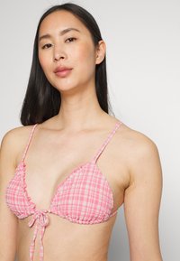 Top de bikini triangular em padrão xadrez rosa com bordas onduladas e laços ajustáveis. Apresenta alças finas e uma textura de tecido leve.