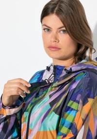 Giacca leggera multicolore con motivi geometrici. Presenta un cappuccio, chiusura con zip e un cordino regolabile per una vestibilità ottimale.
