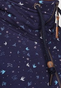 Sudadera azul marino con un cuello alto que presenta un patrón de pájaros y flores. Incluye una cremallera y un cordón con un acento de tope de madera.