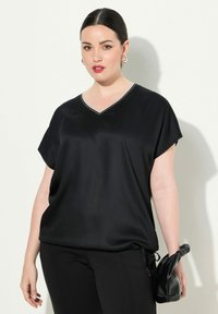 Maglietta nera a V con maniche corte, con una texture liscia simile alla seta, caratterizzata da un motivo decorativo in argento lungo il collo, indossata con pantaloni neri.
