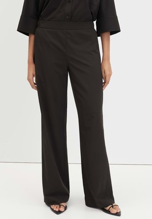 Vrouw met zwarte broek met wijde pijpen en zwarte sandalen, staand tegen een effen lichtgrijze achtergrond.