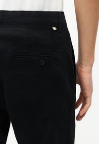 Zbliżenie osoby noszącej czarne spodnie z zapinanym na guzik tylnym kieszonką i małą metalową metką w pobliżu pasa.