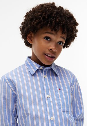 Jeune garçon aux cheveux bouclés portant une chemise à rayures bleues et blanches à boutons, regardant légèrement vers le haut avec une expression pensive.