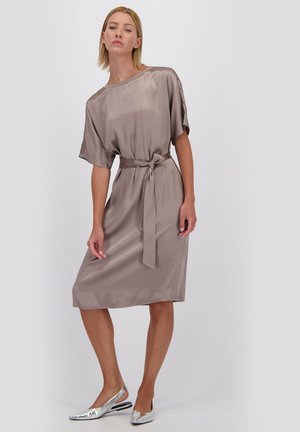 Femme portant une robe midi en satin taupe avec des manches courtes et une ceinture nouée à la taille, associée à des ballerines argentées à talons.
