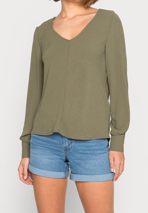 Persoon draagt een olijfgroene blouse met lange mouwen en V-hals, met omgeslagen mouwen en lichtblauwe opgerolde denim shorts.