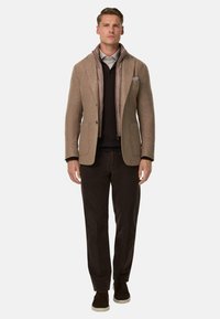 Blazer in misto lana marrone sopra un giubbotto imbottito chiaro con zip, maglione di maglia nera, pantaloni marroni e sneaker scure, il tutto in una palette di colori neutri.