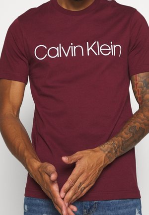 Mężczyzna w burgundowej koszulce Calvin Klein z tatuażami na prawej ręce, trzymający ręce złożone przed sobą.