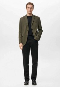 Olivengrøn blazer med hakrevers og enkeltknapslukning, kombineret med en sort sweater og sorte bukser, båret med sorte sko.