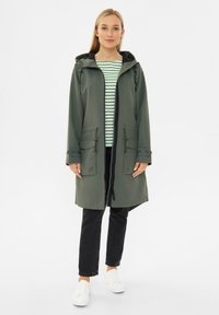 Olijfgroene waterdichte jas met capuchon, aansnoerbare taille, twee voorzakken en ritssluiting, gedragen over een gestreept shirt en zwarte broek.