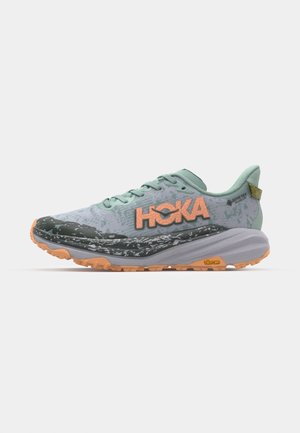 Hoka Trailrunning-Schuh in Hellgrün und Grau, mit strukturierter Oberfläche, auffälligem orangefarbenem Logo und robuster oranger Laufsohle.