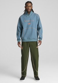 Gaiši zilas džemperis ar oranžiem uzrakstiem un akcentiem, kopā ar olīvzaļām biksēm ar logotipu un kontrastējošiem šuvju elementiem.