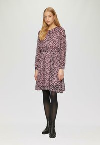 Lila Blumenkleid mit tailliertem Schnitt und langen Ärmeln, das eine durchsichtige Textur aufweist. Kombiniert mit schwarzen Knöchelstiefeln und schwarzen Strumpfhosen.