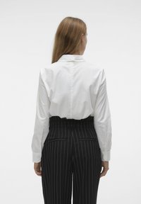 Chemise blanche à manches longues avec col, portée avec un pantalon noir à rayures fines taille haute, doté d'une ceinture et de poches arrière.